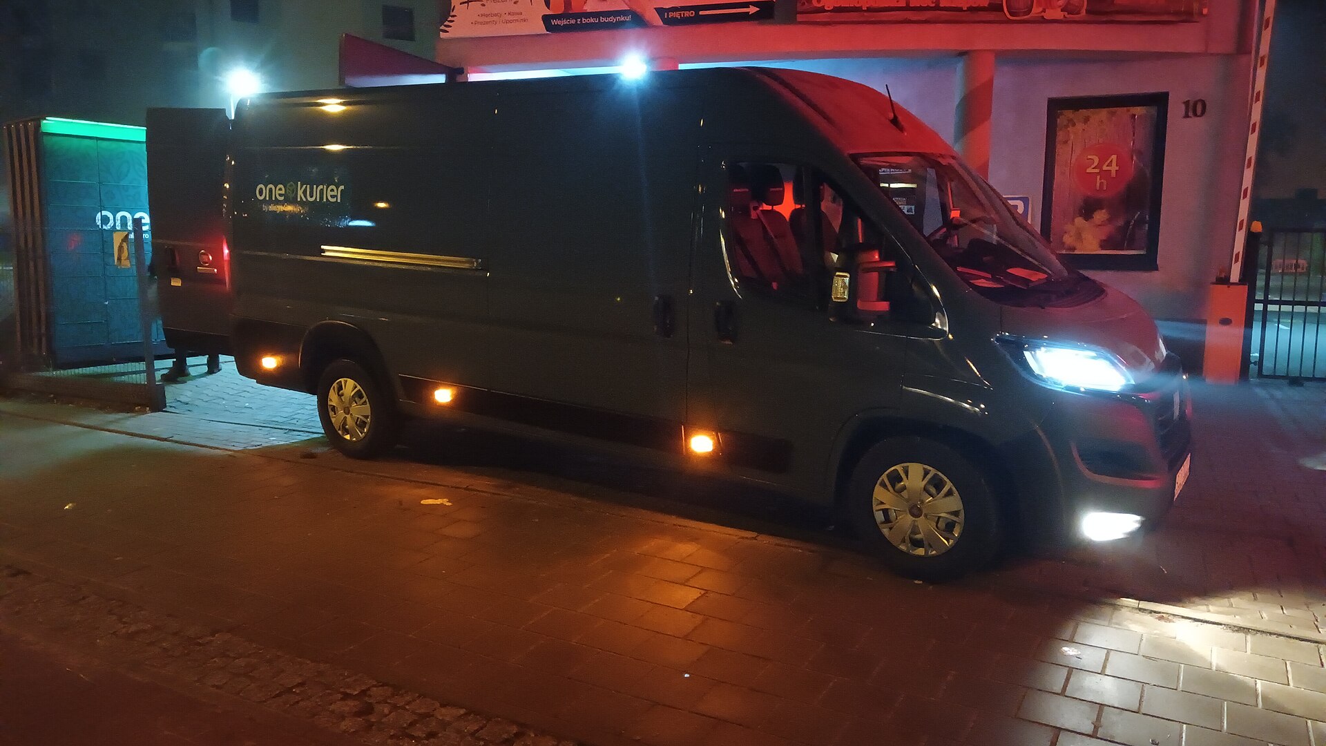 Un utilitar de curierat noaptea, lângă un locker verde în care se livrează colete